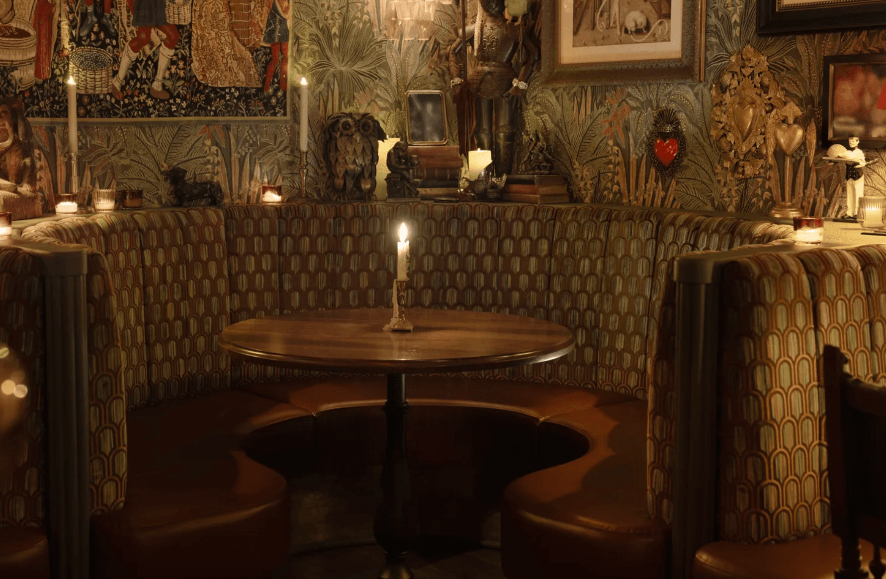 A cozy round booth with a wooden table, a single lit candle, and patterned upholstery sits in a dimly lit room decorated with eclectic art, animal figurines, foliage wallpaper, and playful touches that evoke the charm of Monsieur.